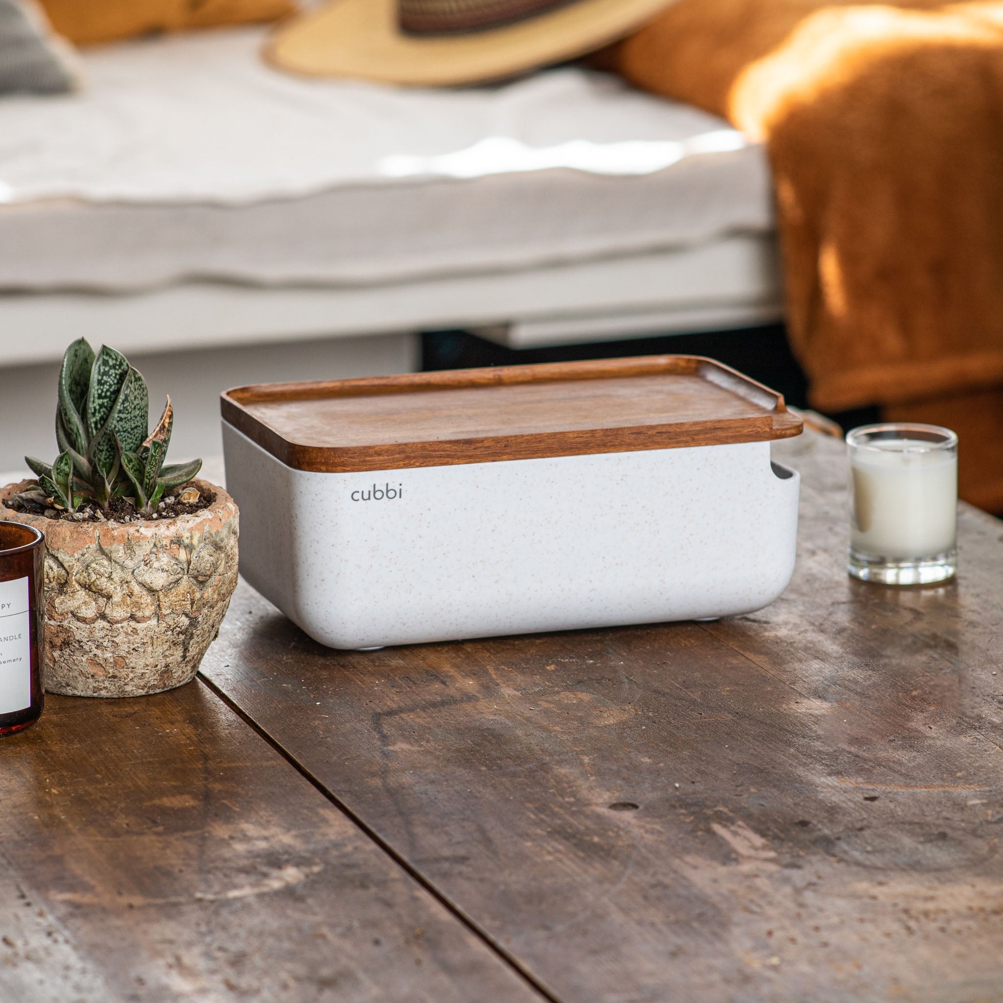 A Cubbi stash box with a wooden lid sits on a top of a wooden table. A small potted plant and a lit candle are positioned to the left of the stash box.