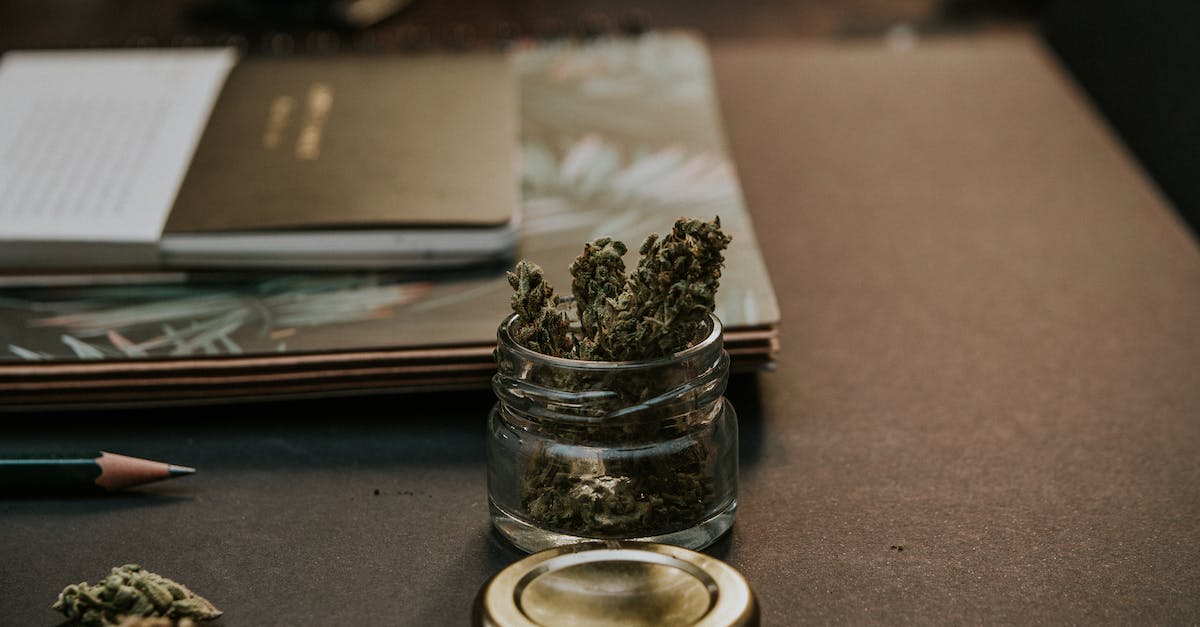 A glass jar containing marijuana sits on a wooden table next to a closed notebook and a pencil.