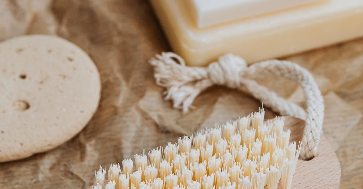 A worn wooden brush with stiff white bristles sits on a scratched wooden table, next to a bar of soap and a smooth, gray rock.